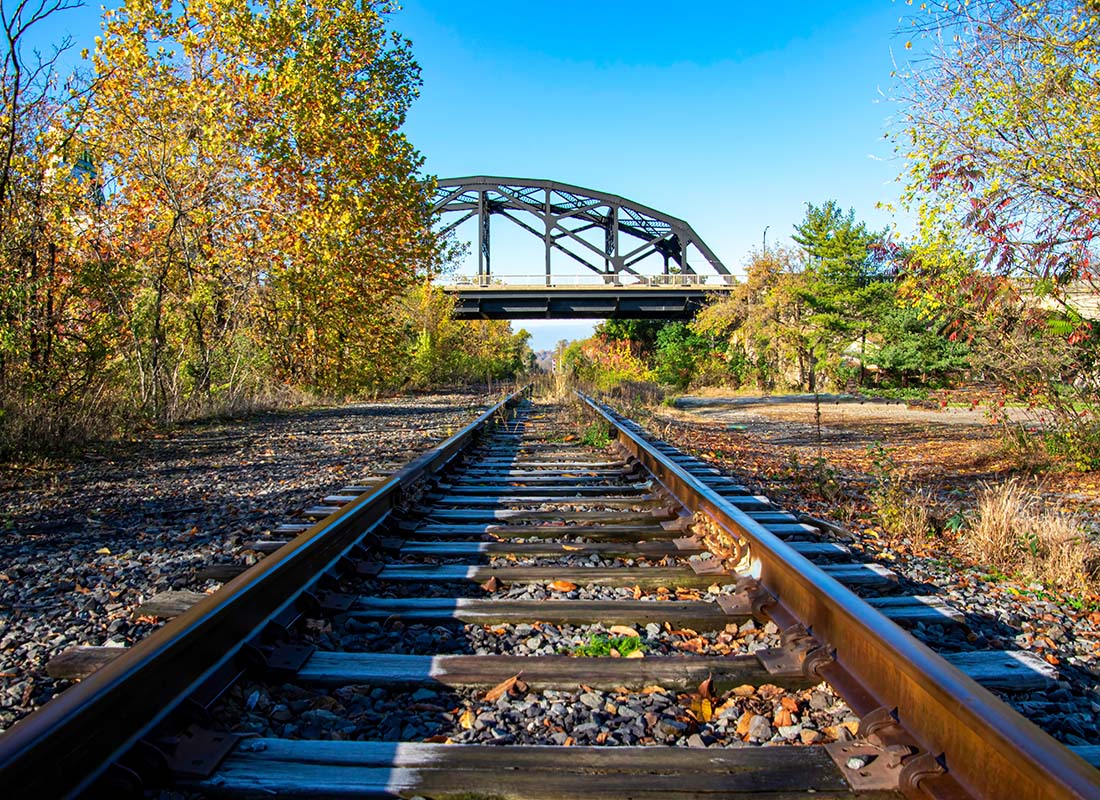 Captive Client Portfolio - Closeup View of Train Tracks in a Secluded Area Surrounded by Trees with a Bridge in the Distance Against a Clear Blue Sky