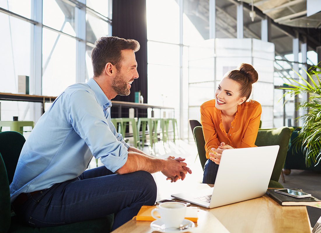 Contact - Portrait of a Cheerful Young Business Man and Woman Sitting Around a Coffee Table with a Laptop and Coffee on Top Having a Meeting in a Brightly Lit Room