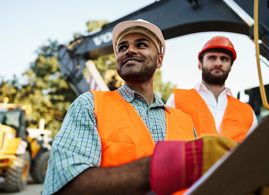 Read Our Reviews - Closeup Portrait of a Smiling Young Contractor Worker Holding a Notepad While Standing Next to his Coworker and Heavy Construction Equipment